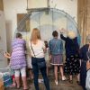 three women are tying coloured string to a mesh circle faintly depicting a sunflower. Each string added will make the image bolder and stronger.