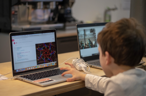 A boy with a white shirt and short, dark brown hair is sat in front of a laptop with his hand on the tracking pad. There is coding on the left of the screen and a kaleidoscope-style image on the right in reds, greens and blues.