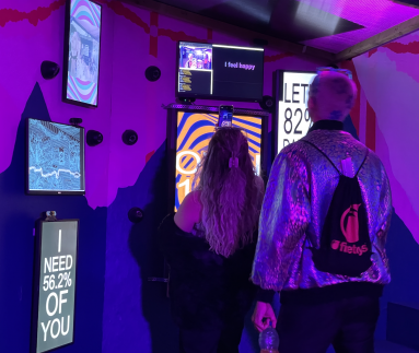 A dark room holds a number of screens, each a different size and presented portrait or landscape. On the screens are brightly coloured waves lines, and one says ‘I need 56.2 percent of you.’ Two people are looking at the screens, one woman with long curly hair in a black jumper and a man with very short blond hair and a silver sequinned jacket.