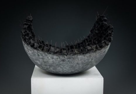 An photograph of a sculpture on a white square plinth. The sculpture is dark black and grey in colour, and is spherical at the bottom, which is smooth and then the sphere is taken away and left with a textured centre.